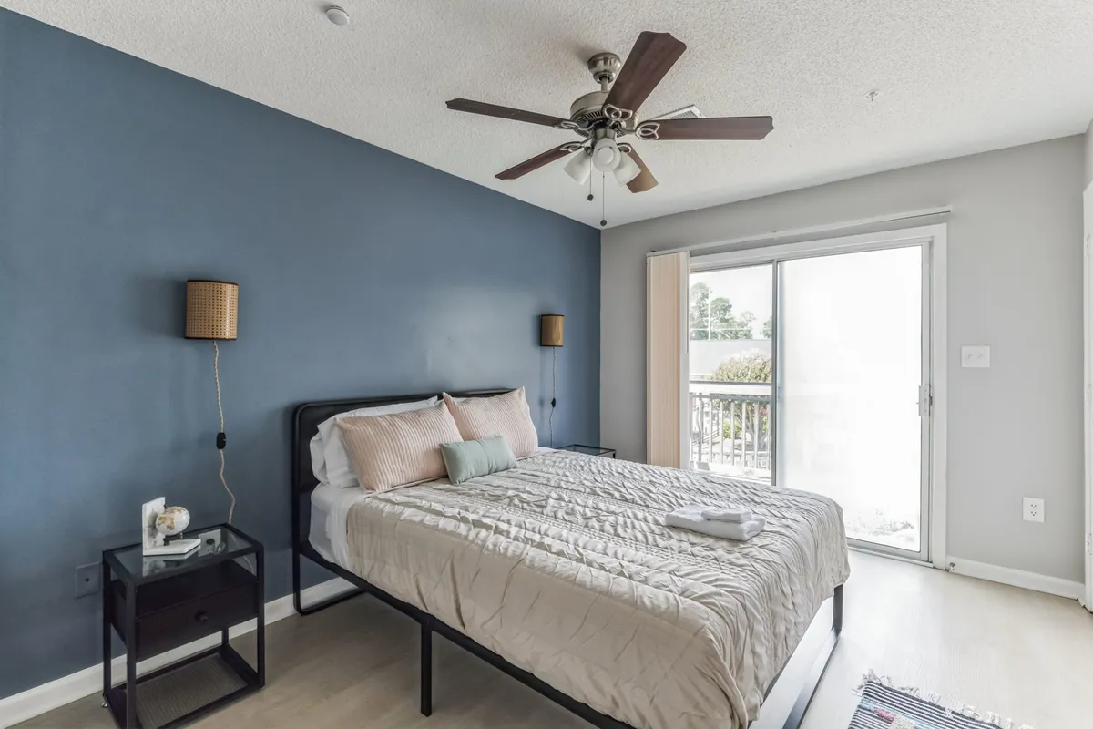 A cozy bedroom highlighted by a large bed, a window letting in warm sunlight, a nightstand, and a ceiling fan above. The decor is understated and soothing, with clean lines and soft tones enhancing the peaceful mood.