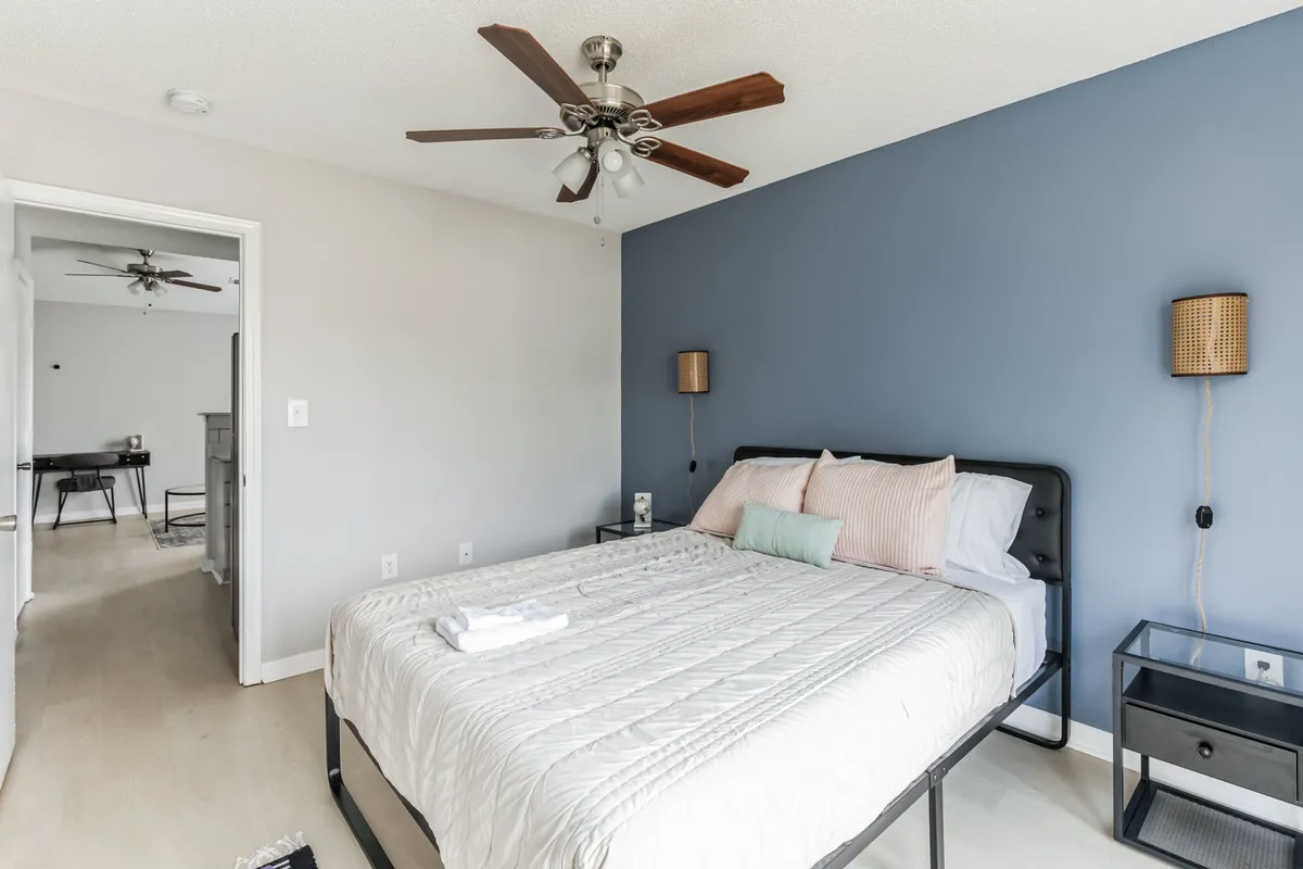 This image presents a serene bedroom scene featuring a bed with light, inviting bedding, complemented by a calming blue accent wall and a nightstand topped with a softly glowing lamp. Gentle natural light fills the room, evoking a peaceful, restful atmosphere ideal for unwinding.
