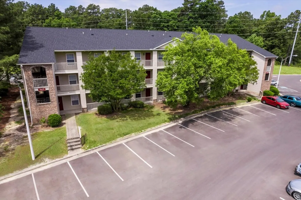 This image shows the welcoming exterior of the apartment building, framed by large trees and a well-maintained parking lot. The setting feels private yet connected to a friendly community, suggesting peaceful strolls or relaxed time spent outdoors together.