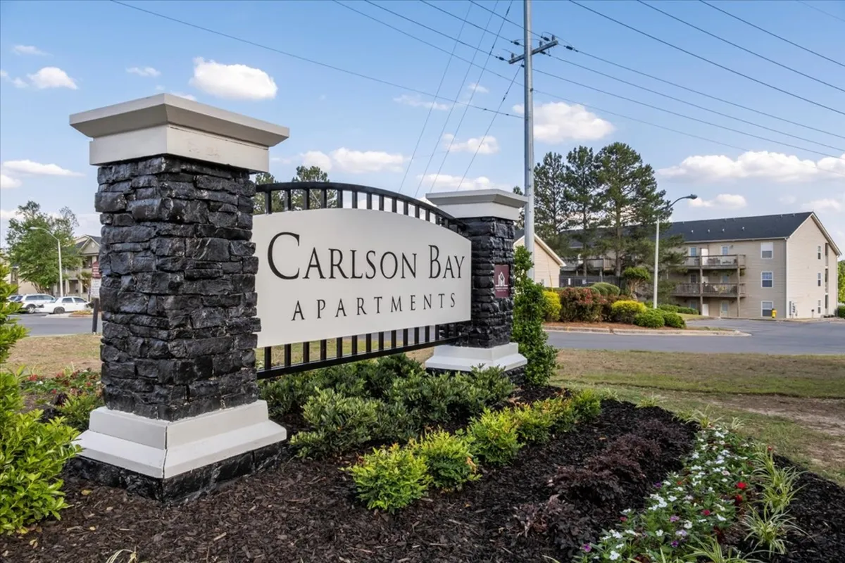 This exterior image features welcoming signage, attractive landscaping, and several surrounding buildings. The scene suggests a vibrant, well-maintained community with easy access to nearby amenities and a sense of arrival.