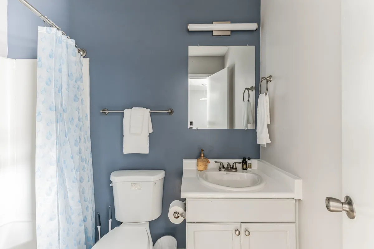 A serene, modern bathroom featuring a clean white sink, a neatly arranged toilet, a mirror reflecting soft natural light, and a simple shower curtain. The setting feels calm and spa-like, ideal for personal grooming or a relaxing bath.