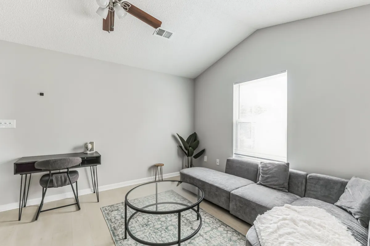 This image presents a modern gray sectional sofa with a glass coffee table atop a decorative area rug. A desk with a chair is visible in the corner, suggesting the space can be used for both work and relaxation. The overall mood is serene and inviting.