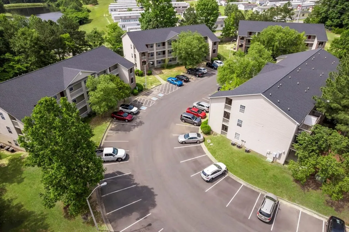 An exterior view of a residential area with a parking lot framed by lush greenery. Nearby, families and neighbors gather for outdoor activities and casual meetups, set against a backdrop of residential buildings and mature trees.