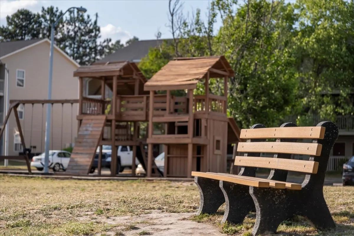 A vibrant exterior scene featuring a playground structure and a park bench, surrounded by families and children enjoying the outdoors. The area is filled with greenery, creating a lively and inviting atmosphere perfect for recreation and community bonding.