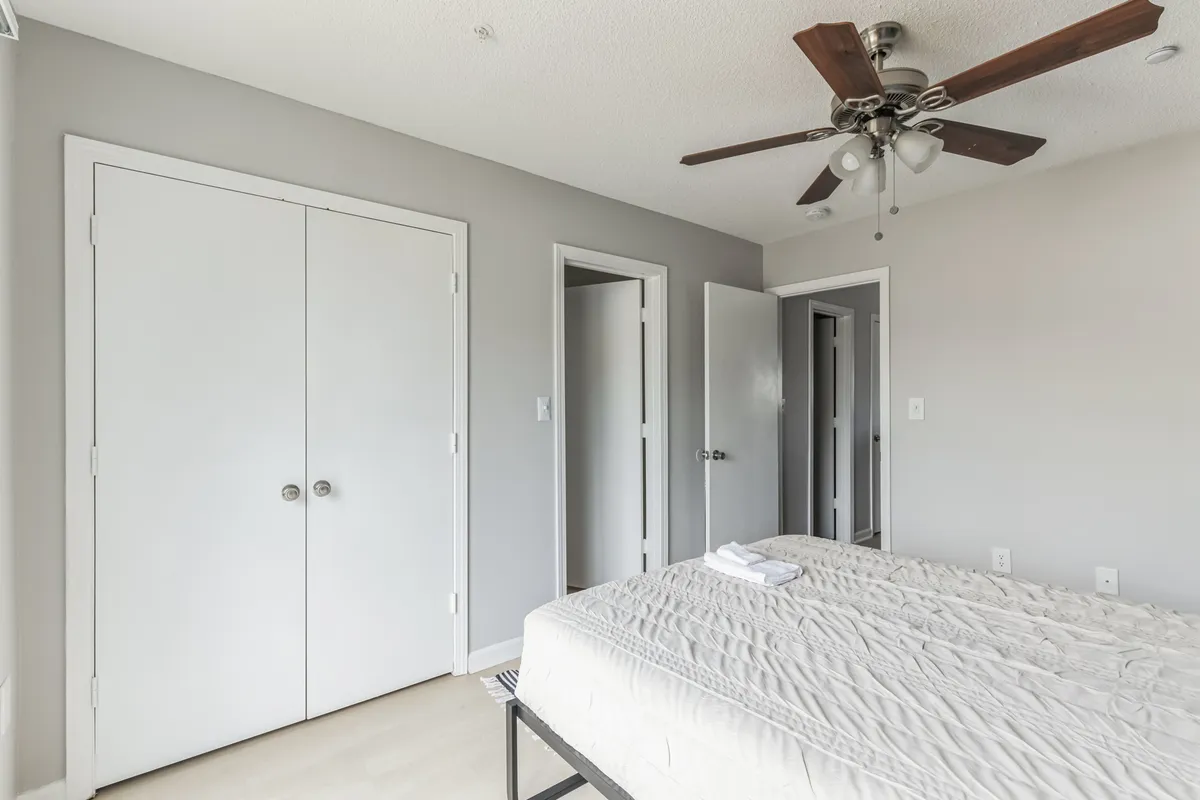 A spacious bedroom featuring a comfortable bed, ceiling fan, and double closet doors. The room is uncluttered and airy, with space for relaxing, reading, or preparing for the day, reinforcing the suite’s versatility and home-away-from-home feel.