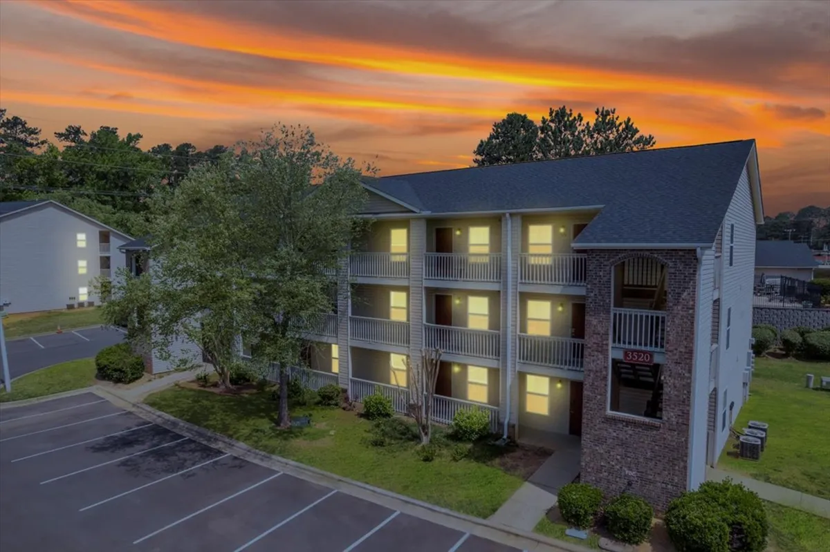 The image showcases the exterior of a modern apartment building at sunset, with warm light glowing from the windows. A balcony offers a perfect spot to relax and take in the tranquil evening sky, while the surrounding area feels peaceful and inviting.